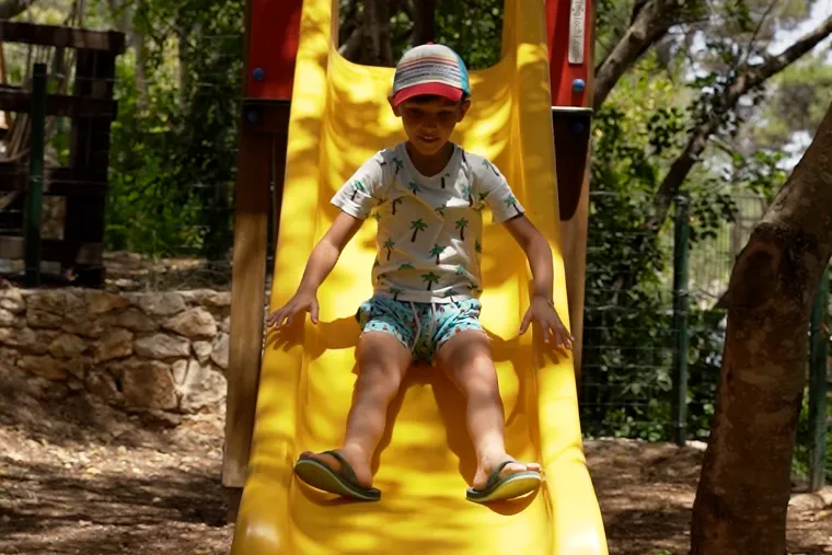 Bambino sullo scivolo dell'area giochi, tra i pini